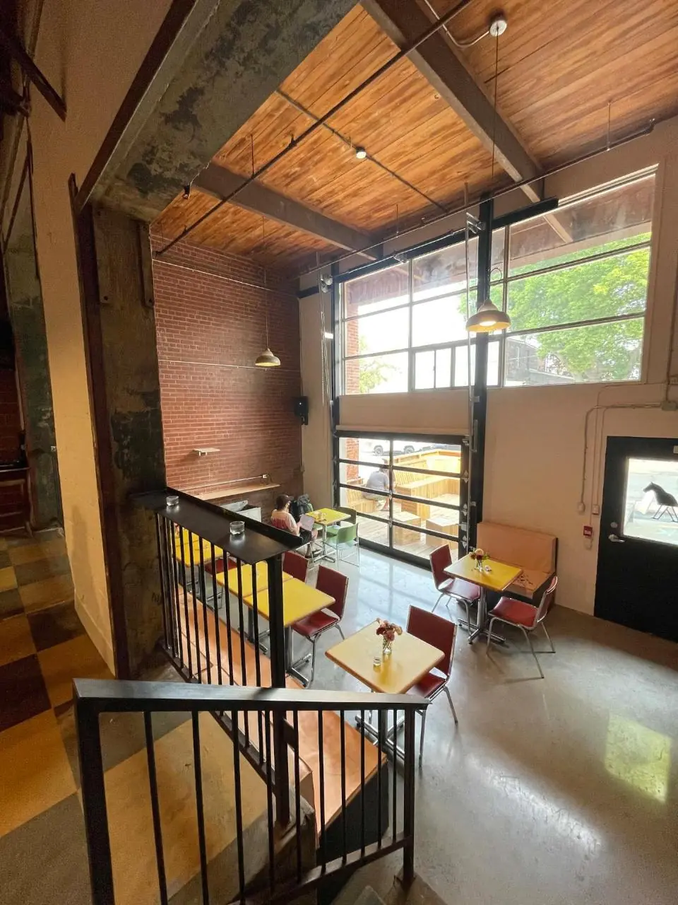 The main floor of Vers seen from the mezzanine stairs: wood plank ceiling, brick wall, tall factory windows, yellow tables and red chairs
